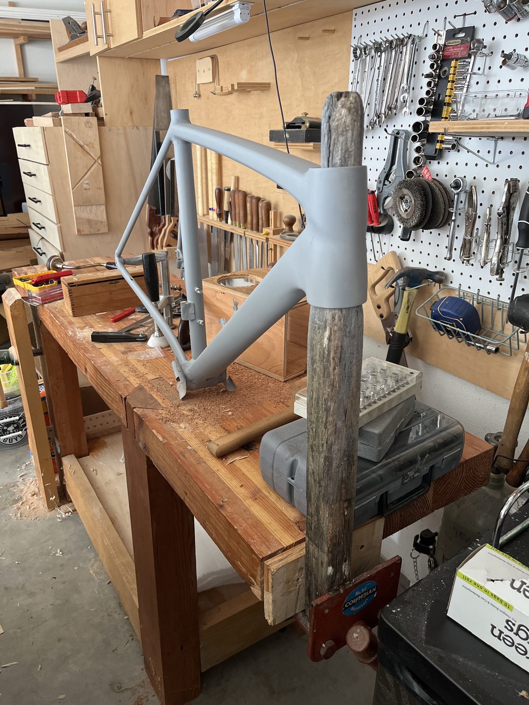 A bare bicycle frame clamped in a repair stand in front of a pegboard of tools.