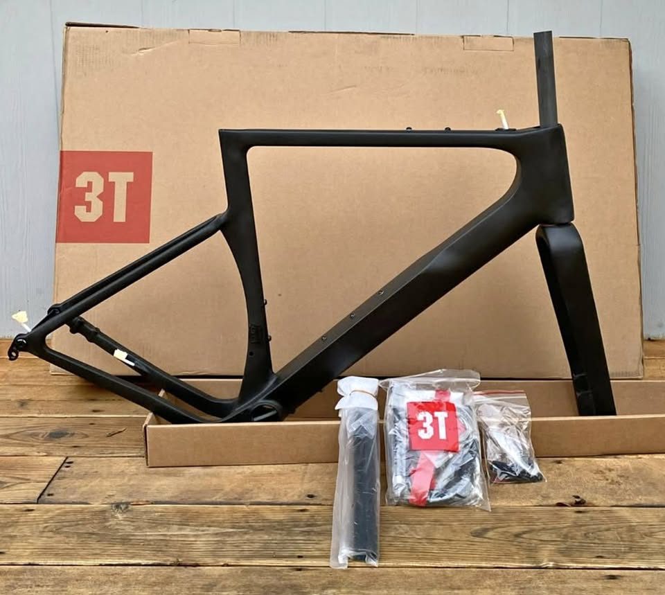 Unpainted black carbon 3T frame and fork standing in front of the 3T cardboard box they came in, with a wrapped seatpost and two sealed bags of hardware on the boards in front, from the seller's Marketplace listing.