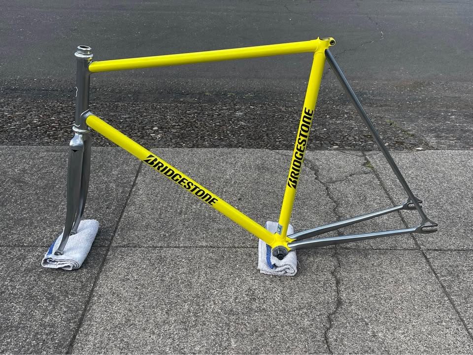 Yellow Bridgestone track frame with a polished silver rear triangle propped on a pavement beside its matching silver fork, from the seller's Marketplace listing.