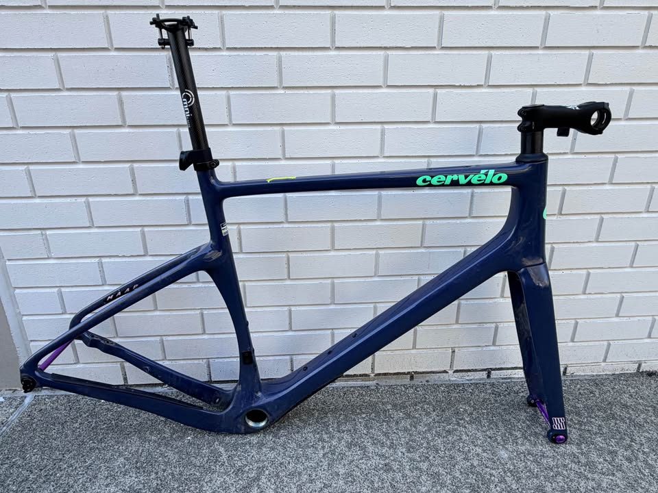 Dark blue Cervélo Áspero frame and fork with a stem and seatpost fitted and purple axles, leaning against a white brick wall, from the seller's Marketplace listing.