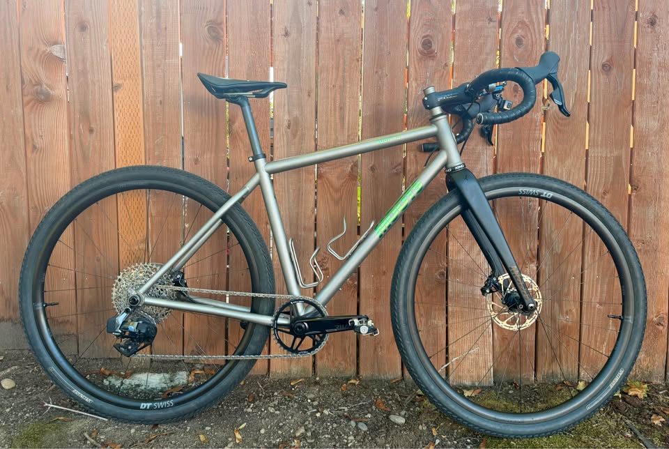Titanium Moots Routt 45 built up as a complete bike with green Moots graphics, a black carbon fork and DT Swiss wheels, leaning against a wooden fence, from the seller's Marketplace listing.