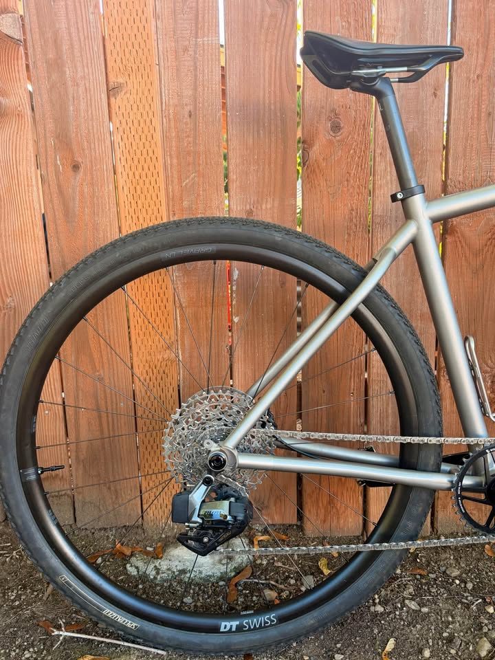 Titanium Moots Routt 45 built up as a complete bike with green Moots graphics, a black carbon fork and DT Swiss wheels, leaning against a wooden fence, from the seller's Marketplace listing. Photo 2 of 8.