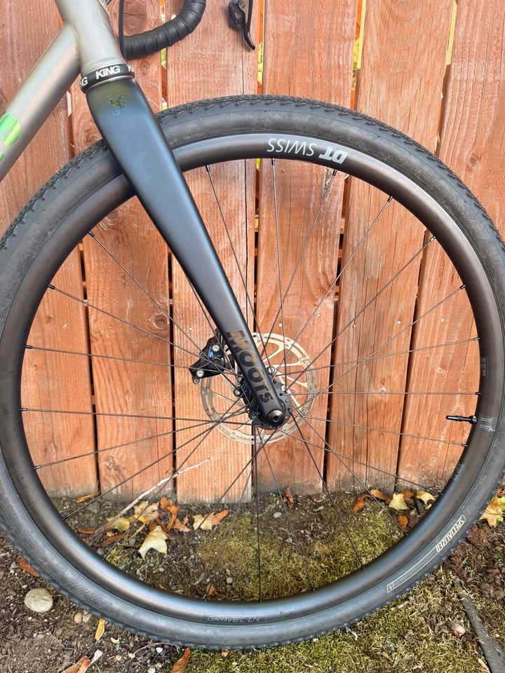 Titanium Moots Routt 45 built up as a complete bike with green Moots graphics, a black carbon fork and DT Swiss wheels, leaning against a wooden fence, from the seller's Marketplace listing. Photo 4 of 8.