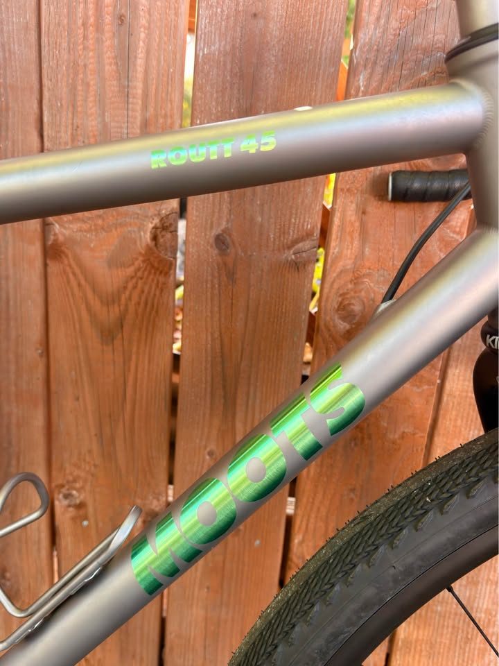 Titanium Moots Routt 45 built up as a complete bike with green Moots graphics, a black carbon fork and DT Swiss wheels, leaning against a wooden fence, from the seller's Marketplace listing. Photo 6 of 8.