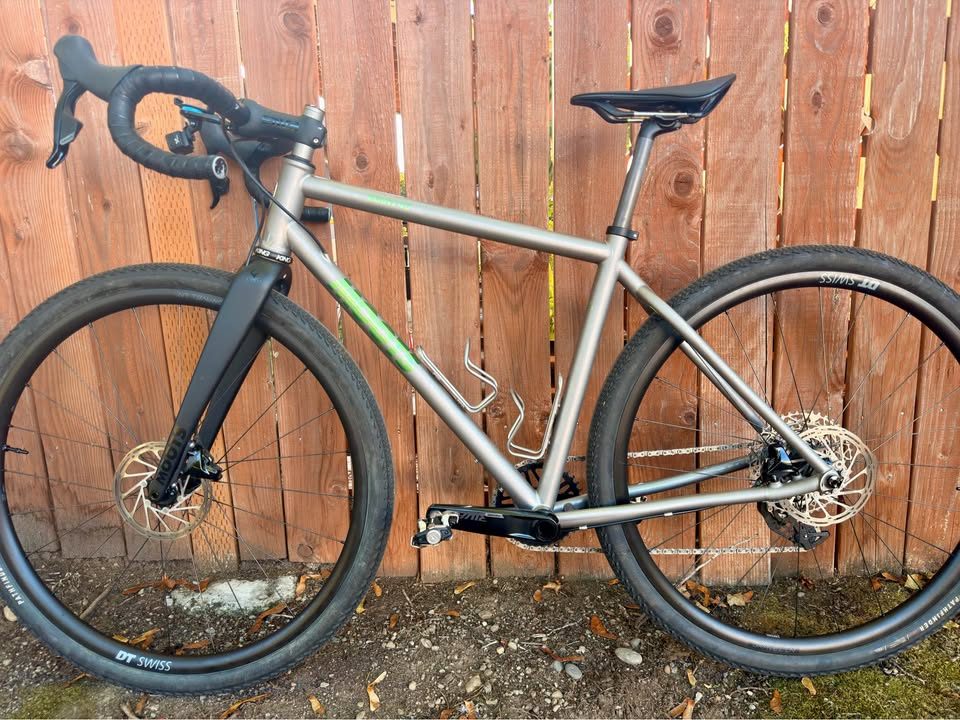 Titanium Moots Routt 45 built up as a complete bike with green Moots graphics, a black carbon fork and DT Swiss wheels, leaning against a wooden fence, from the seller's Marketplace listing. Photo 8 of 8.