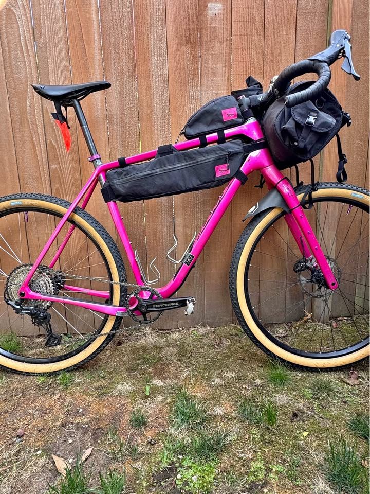 Hot pink Salsa Cutthroat leaning against a wood fence, built up with drop bars, tan wall knobby tyres and a frame bag and seat pack, from the seller's Marketplace listing.
